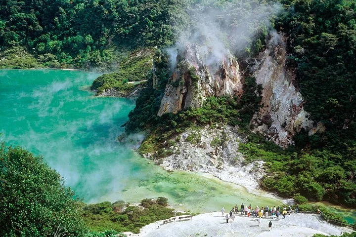 Eine Gruppe von Touristen hat sich in der Nähe eines dampfenden grünen Thermalsees und felsiger Klippen versammelt.