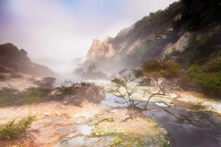 Een mistig, desolaat landschap met een kleine stroom die door kleurrijke geothermische afzettingen stroomt.
