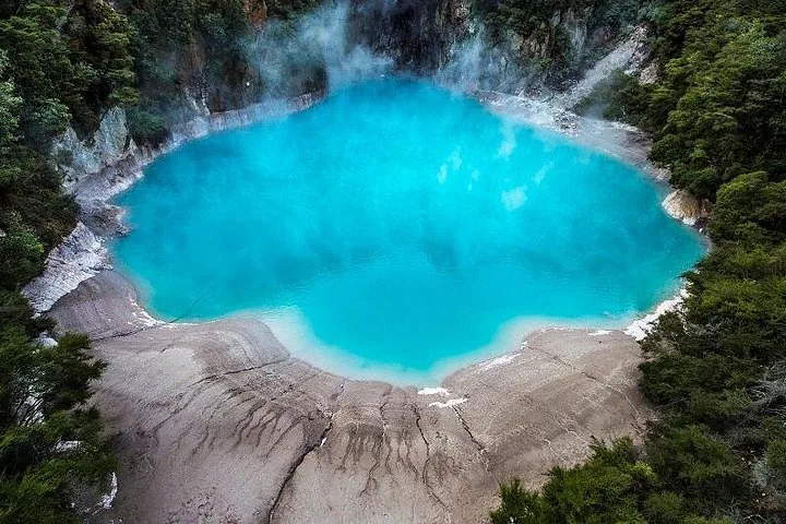Hellblaue heiße Quelle, die Dampf ausstößt, eingefasst von grauer Kruste und grüner Vegetation.