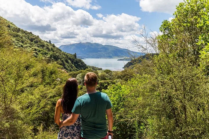 Paar betrachtet von einer grünen, bewaldeten Aussichtsplattform aus eine Seen- und Berglandschaft.