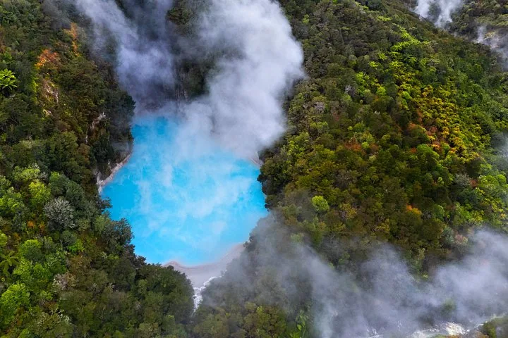 Aerial view of a bright blue steaming geothermal pool surrounded by forest